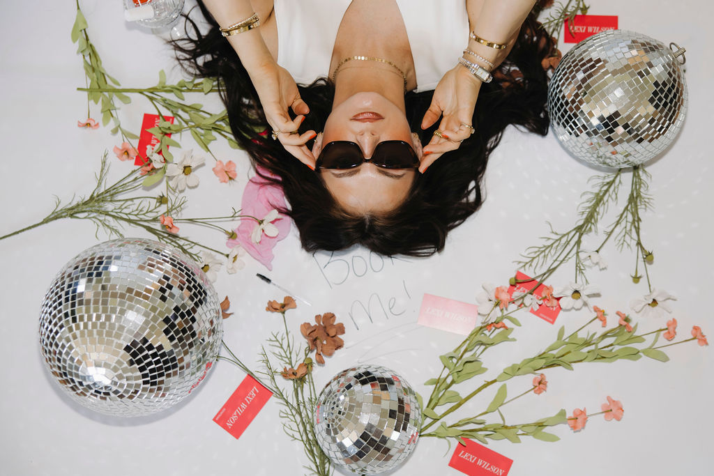 Louisville branding photographer | an esthetician laying on the floor on top of a piece of paper that says 'book me!' and flowers and disco balls around her.