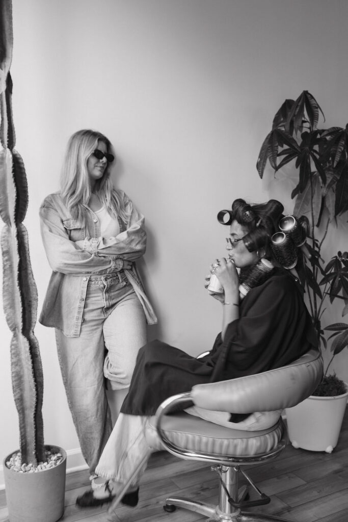Black and white photo of a hair client wearing foils, sunglasses while the hairstylist stands next to her.