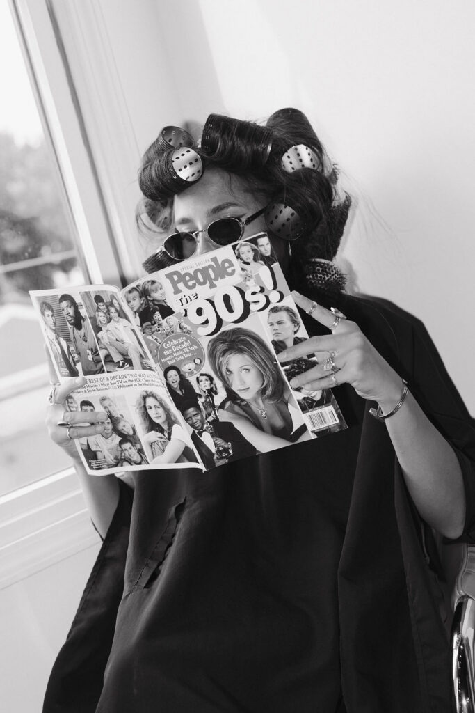 Black-and-white portrait of a hairstylist wearing rollers and sunglasses while reading a 1990s magazine during her branding session.