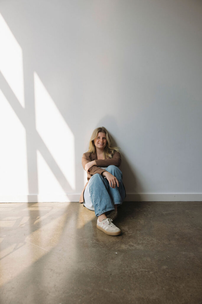Casual professional portrait of a Louisville hairstylist sitting against a sunlit studio wall.