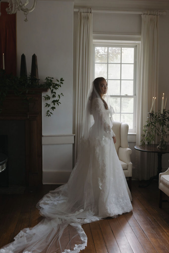 Bride in a lace wedding gown with a long train standing beside a window at Hermitage Farm.