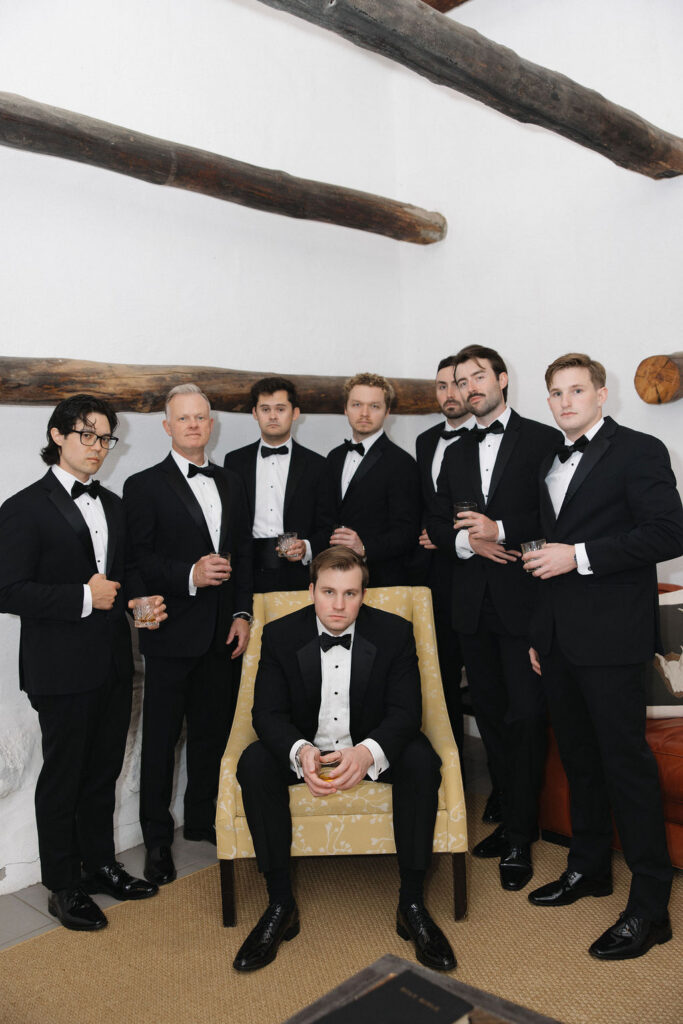 Groom seated in a yellow armchair surrounded by groomsmen in black tuxedos at Hermitage Farm.