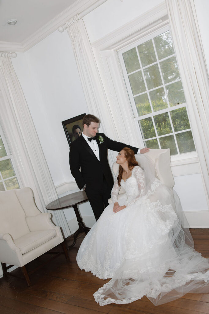 Bride seated with her gown and veil spread across the floor as a man stands behind her