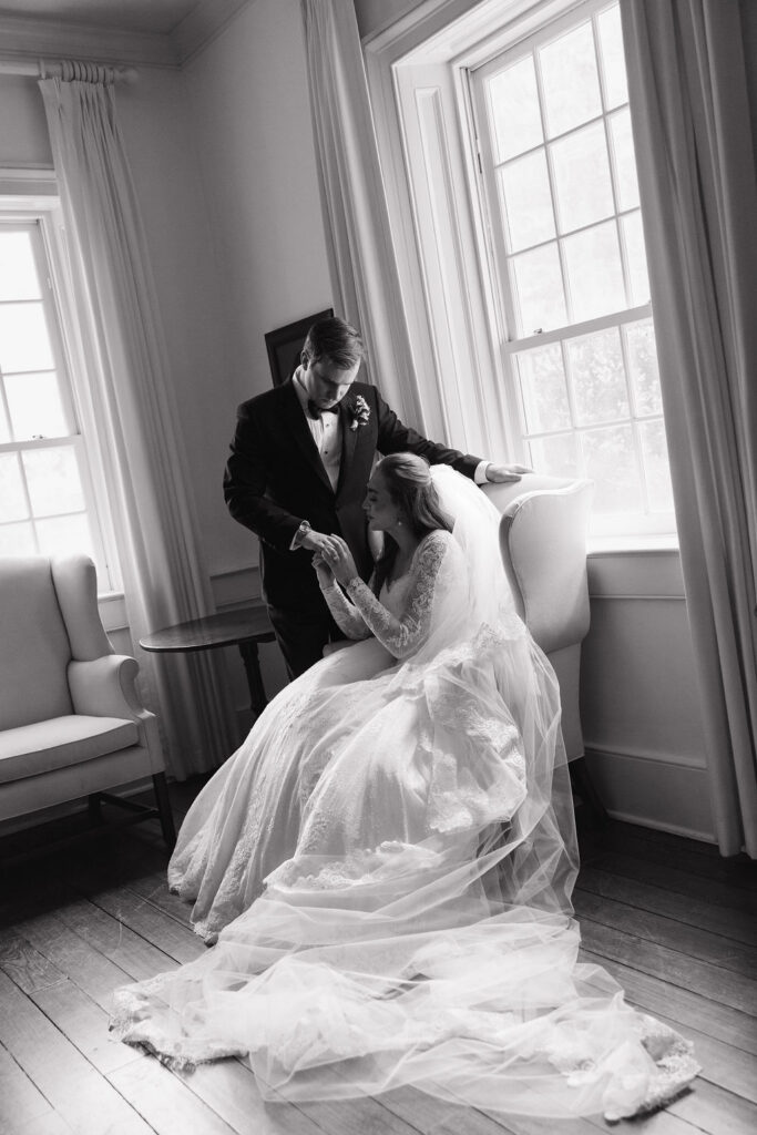 Bride seated with her gown and veil spread across the floor as a man stands behind her in black and white photo at a black tie wedding at Hermitage Farm.