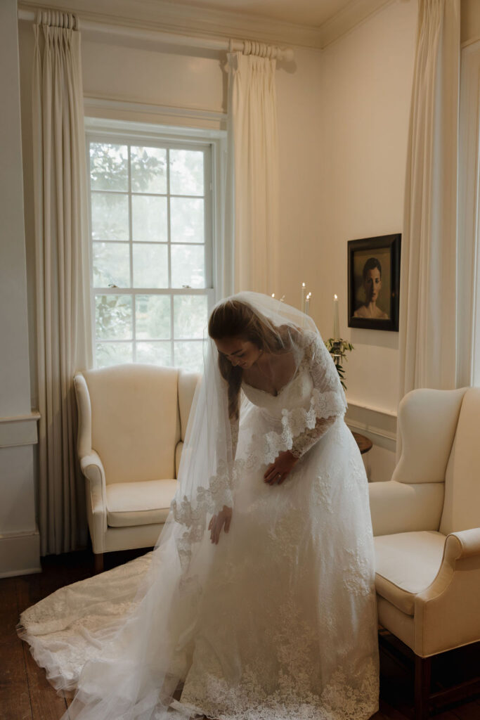 Bride adjusting her lace wedding gown beside an upholstered armchair