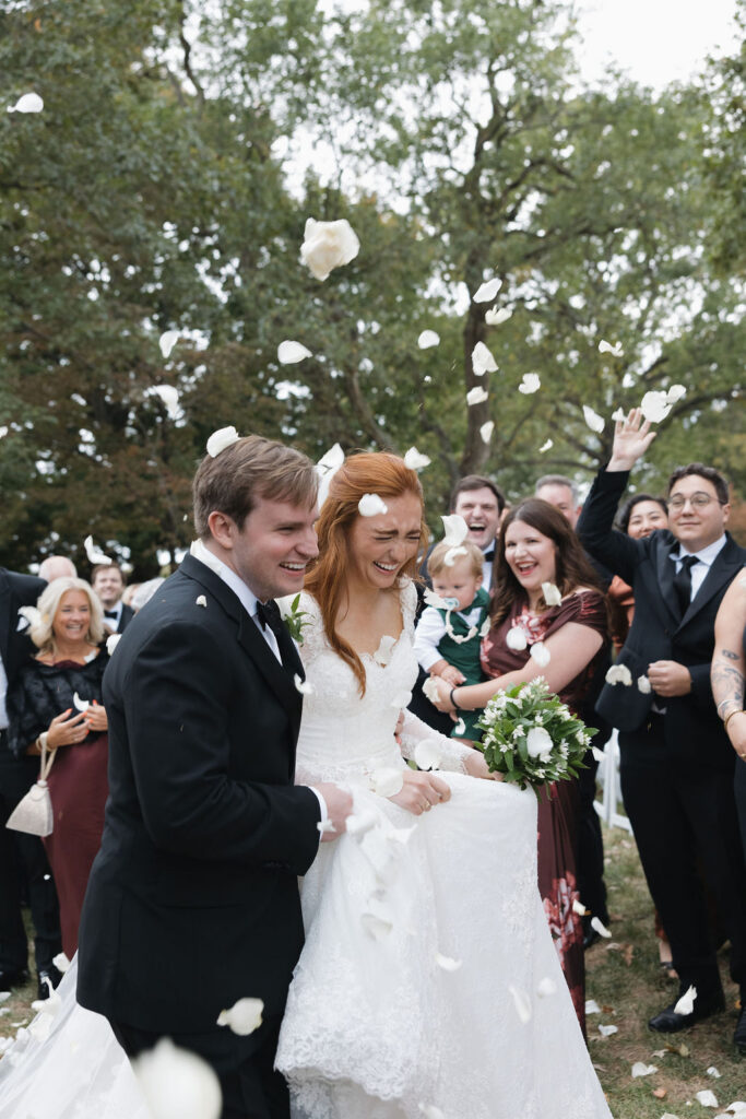 Newlyweds smiling as guests toss white petals around them at a black tie wedding at Hermitage Farm.