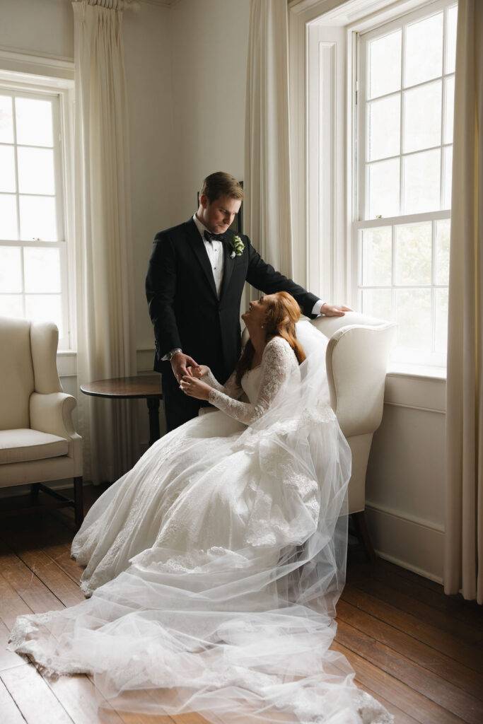 Bride seated with her gown and veil spread across the floor as a man stands behind her at their black tie wedding at Hermitage Farm.